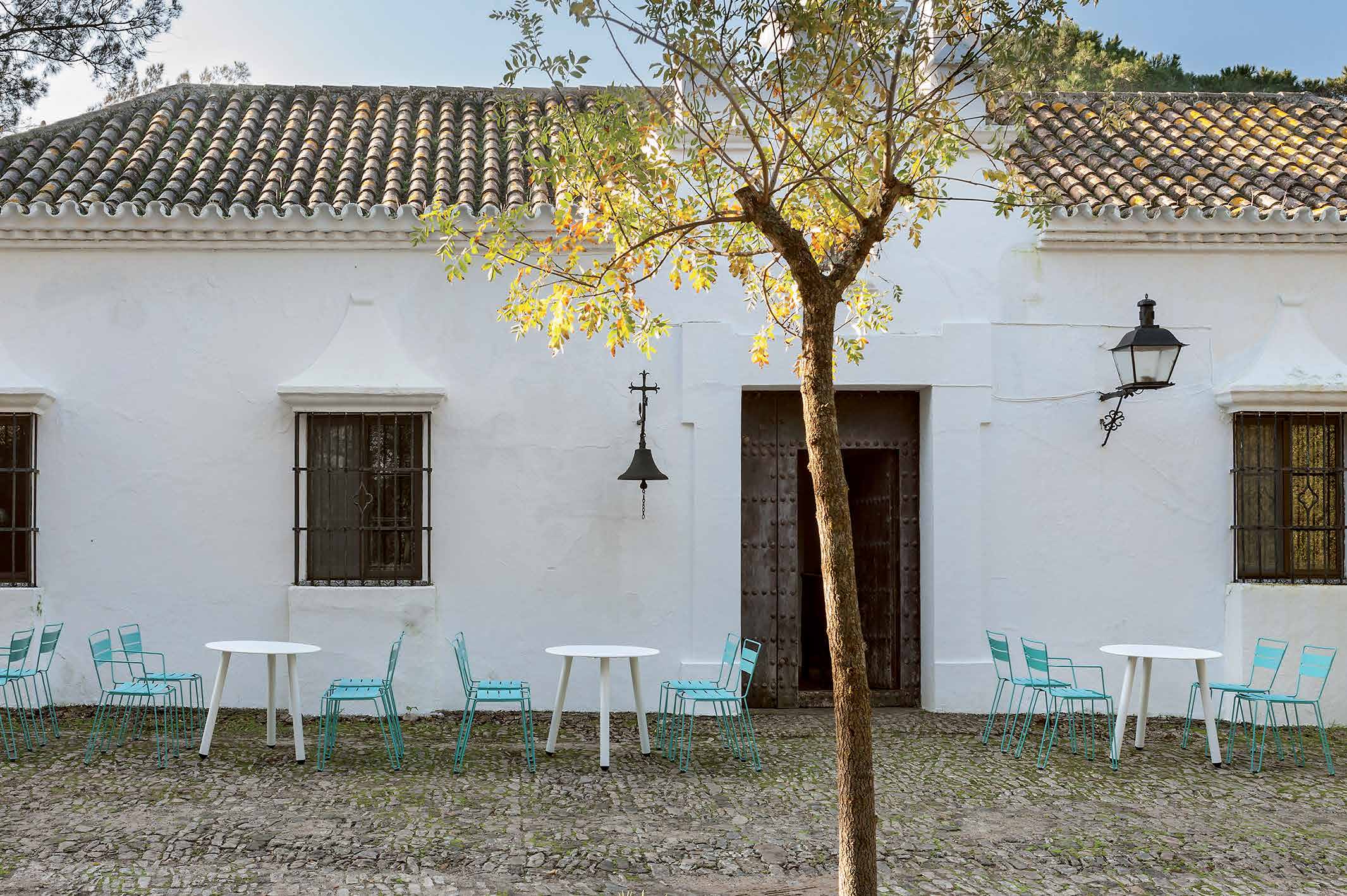 Turquoise café chairs and round white tables outside a whitewashed Andalusian farmhouse, under a terracotta tiled roof — iSiMAR catalogue photography