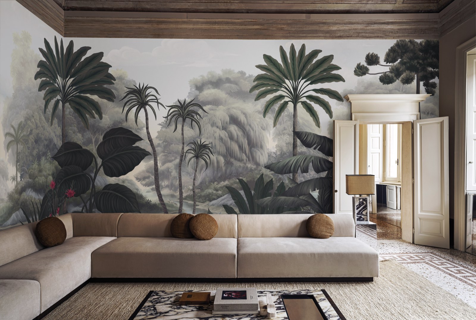 A wide grey-green jungle mural — palms, banana leaves and a weeping willow above a pale sky — covering the long wall of a room with a coffered timber ceiling, a low cream sectional sofa and a terrazzo floor.