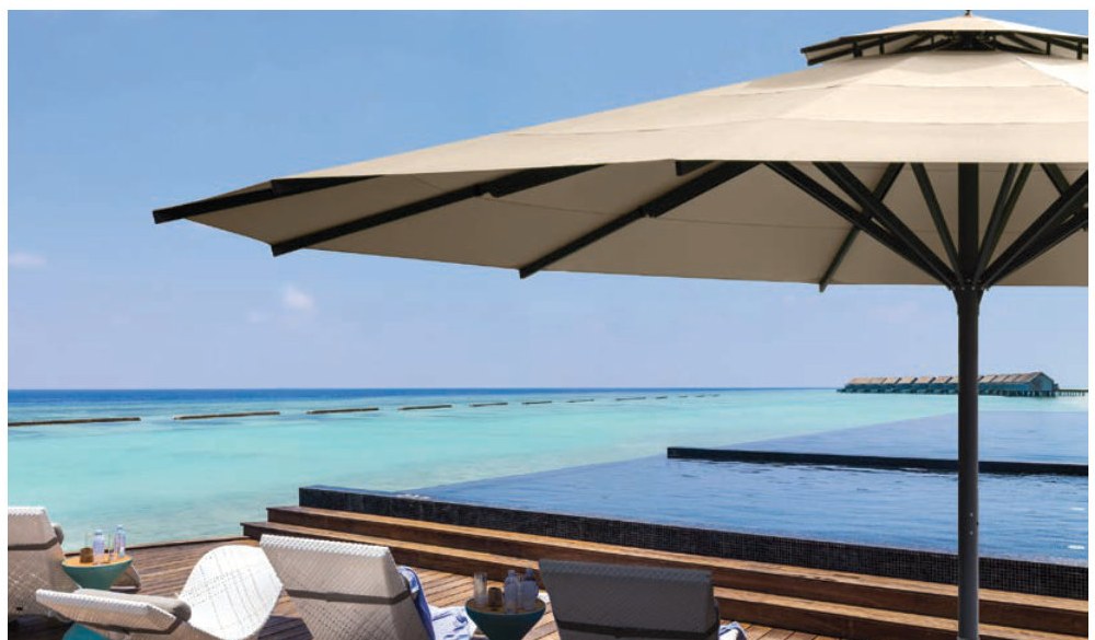 Large round cream cantilever parasol over an overwater deck, turquoise lagoon and stilted bungalows beyond